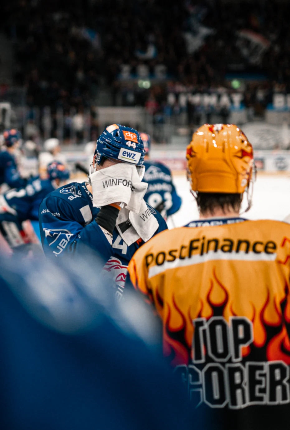 Erschöpfter Eishockeyspieler in blauem Trikot wischt sich mit Handtuch das Gesicht, neben einem Teamkollegen im orange-gelben Top-Scorer-Trikot. Die Szene zeigt die Intensität eines Matches in einer belebten Eishockeyarena.