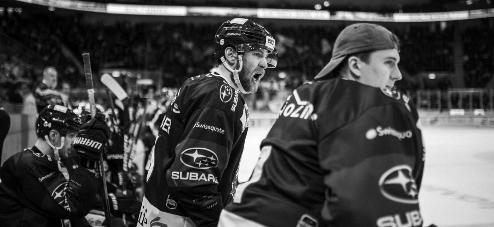 Emotionaler Eishockeyspieler, Nummer 6, mit Bart und Helm, ruft auf der Spielerbank im schwarz-weißen Bild. Ein Teamkollege sitzt daneben, Zuschauer im Hintergrund. Trikots zeigen Sponsorenlogos wie "Subaru" und "Swissquote".
