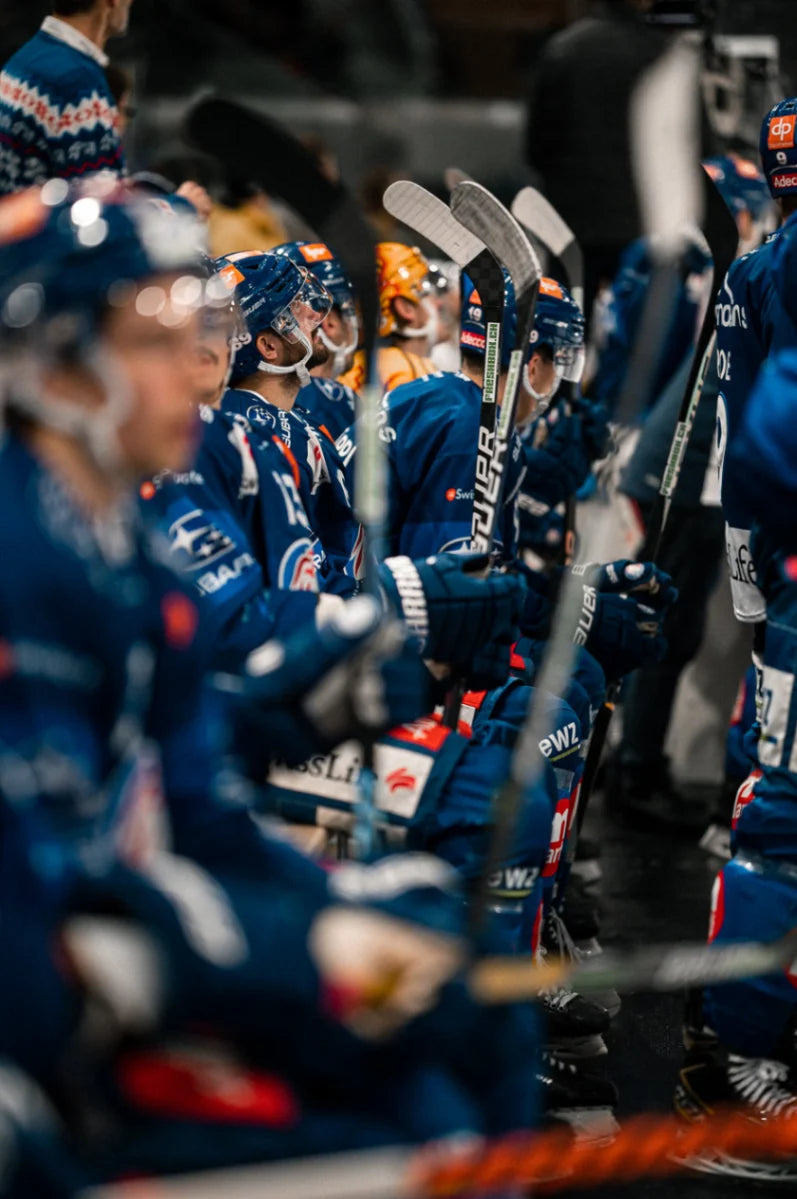 Eishockeyspieler in blauen Trikots, Helmen und mit Schlägern sitzen konzentriert auf der Ersatzbank. Die professionelle Ausrüstung und sichtbare Sponsorenlogos betonen die Wettkampfatmosphäre vor dem nächsten Einsatz auf dem Eis.