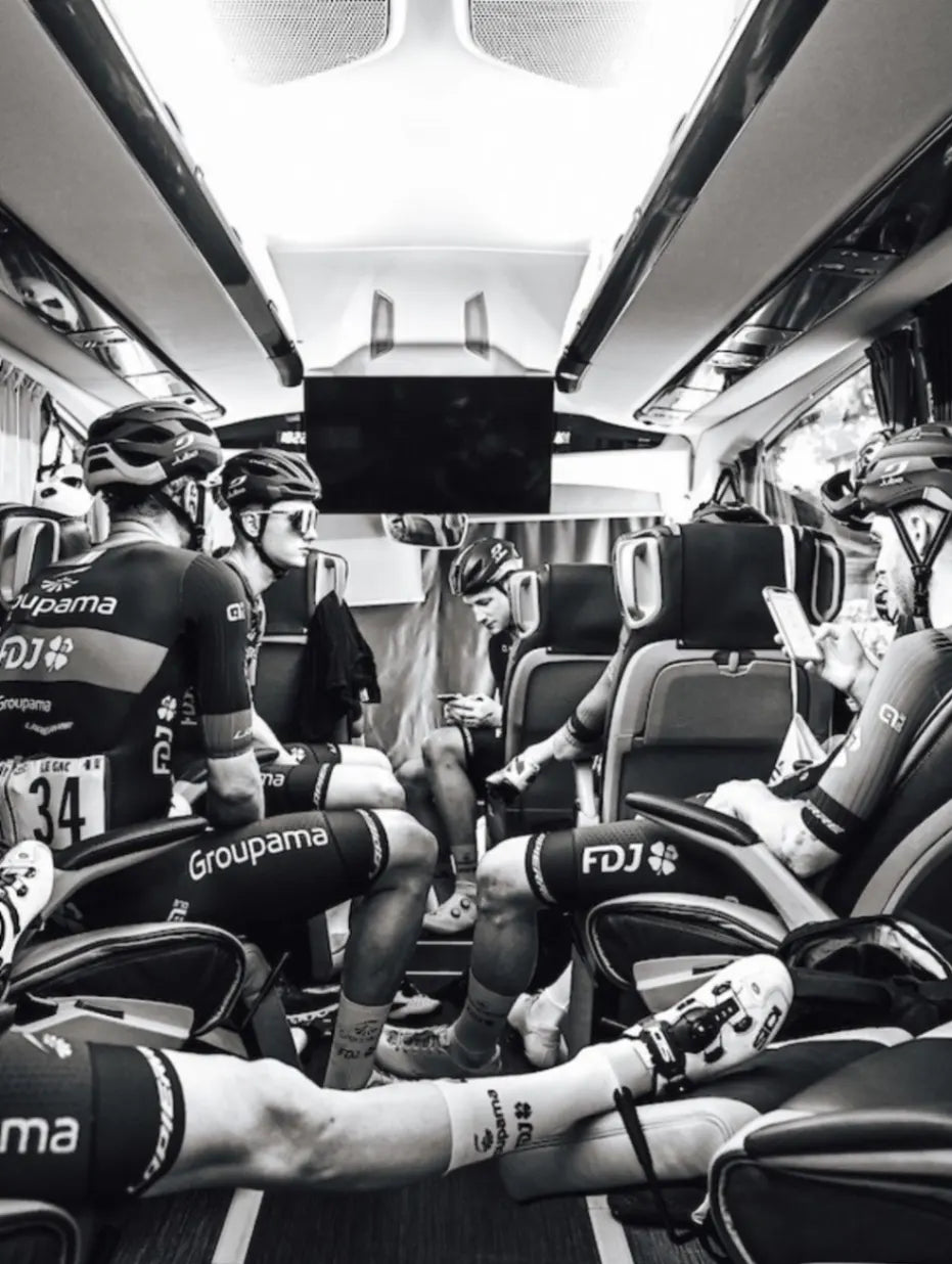Schwarz-Weiß-Aufnahme professioneller Groupama-FDJ Radfahrer in ihren Teamtrikots und Helmen, die im modernen Teambus entspannen. Sie nutzen Handys und erholen sich vor oder nach dem Wettbewerb.