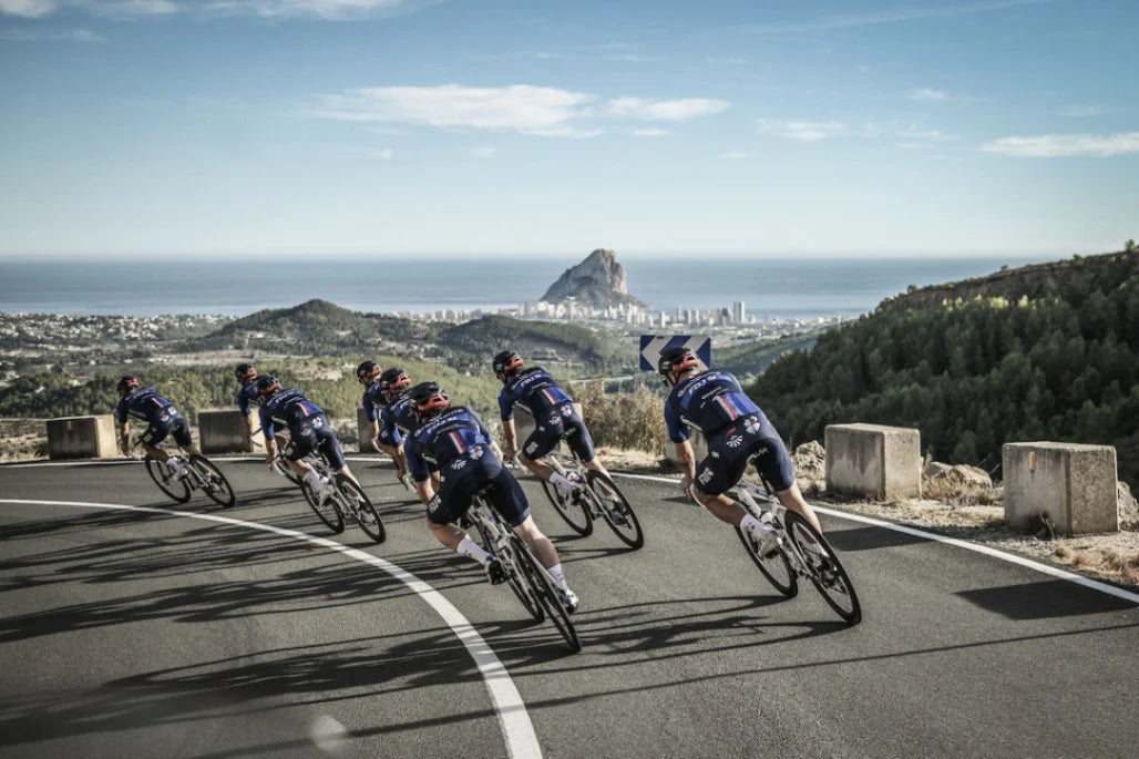 Profiradfahrer des Teams Groupama-FDJ fahren in einer Gruppe eine kurvige Bergstraße hinab, mit der malerischen Küstenstadt Calpe, dem markanten Penon de Ifach Felsen und dem weiten Mittelmeer im sonnigen Hintergrund.
