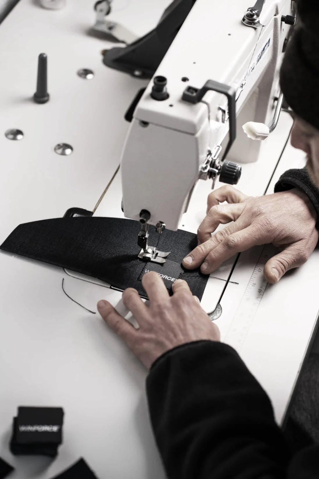 Nahaufnahme eines erfahrenen Schneiders, der mit präzisen Bewegungen schwarzen Stoff unter dem Nähfuß einer professionellen Industrienähmaschine führt. Das sichtbar angebrachte 'WINFORCE' Label deutet auf sorgfältige Textilproduktion und Handarbeit mit Fokus auf Qualität hin.