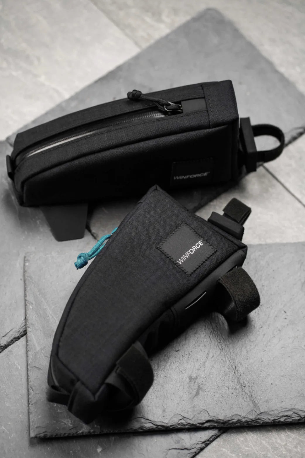 Zwei hochwertige schwarze Winforce Fahrradtaschen liegen auf einer dunklen, strukturierten Schieferoberfläche. Die vordere ist eine kompakte Rahmentasche mit Klettverschlüssen, die hintere eine längliche Oberrohrtasche mit Reißverschluss für Gravel- oder Mountainbikes.