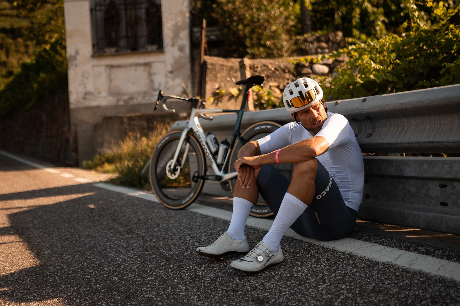Ein erschöpfter Radfahrer in weißem Rennrad-Outfit, Helm und Sonnenbrille sitzt mit seinem Cannondale Rennrad am Straßenrand, eine Pause machend auf einer malerischen Bergstraße in der goldenen Stunde.