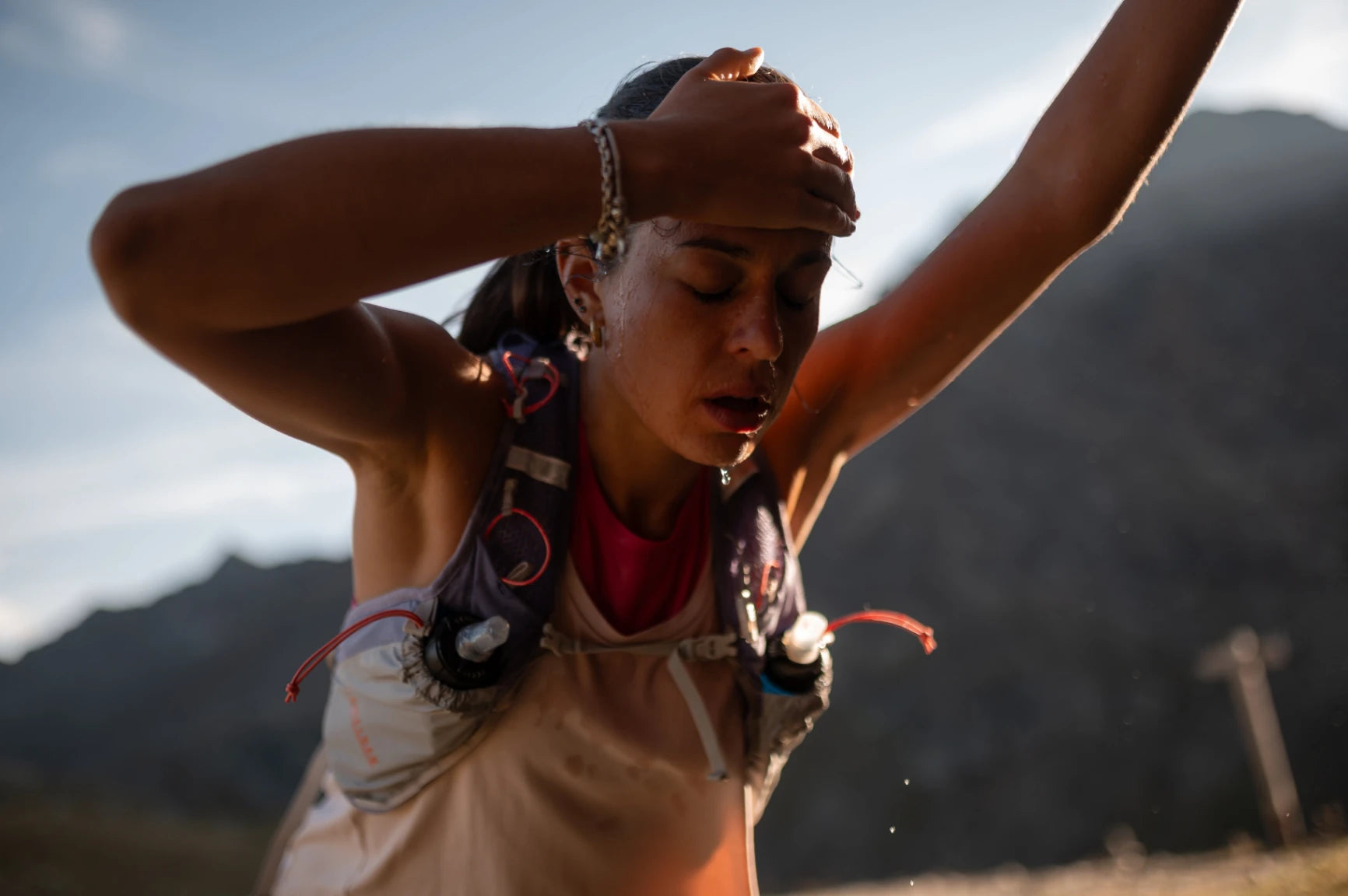 Erschöpfte Trailrunnerin mit verschwitztem Gesicht, die Stirn haltend und mit Wasser vom Kinn tropfend. Sie trägt eine Trinkweste in heller Bergluft, symbolisierend extreme Ausdauer beim Berglauf.