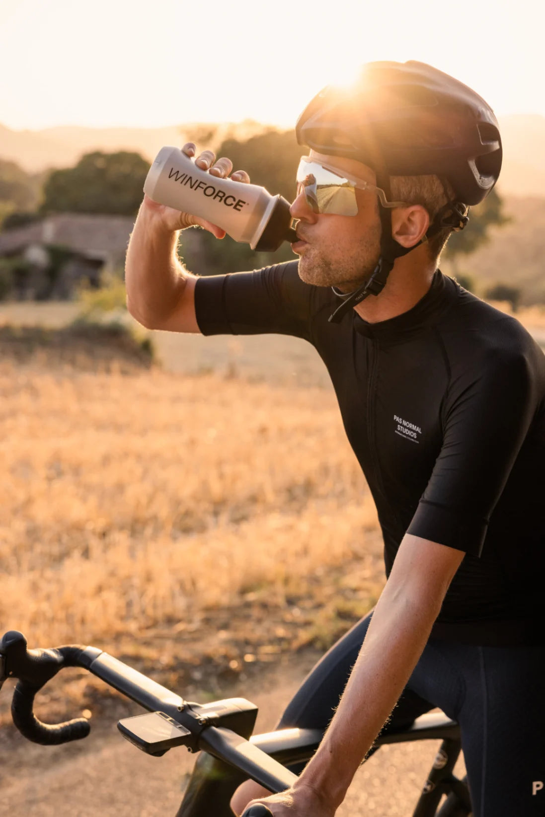 Ein Radfahrer in schwarzem Pas Normal Studios Trikot macht eine Trinkpause aus einer grauen Winforce-Flasche. Die goldene Abendsonne blendet von hinten und taucht die Szene in warmes Licht, während er in einem Feld steht.