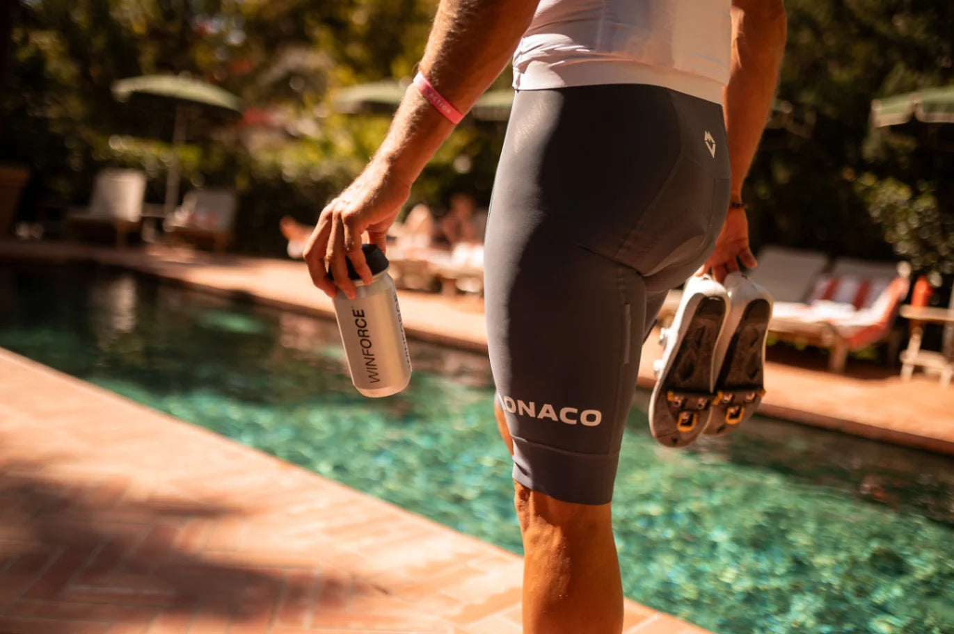 Nahaufnahme eines Radfahrers in grauen Radshorts mit 'MONACO'-Aufschrift, der eine Winforce-Trinkflasche und Fahrradschuhe am Poolrand hält. Die sonnige Szene vermittelt Erholung und Hydration nach dem Training.